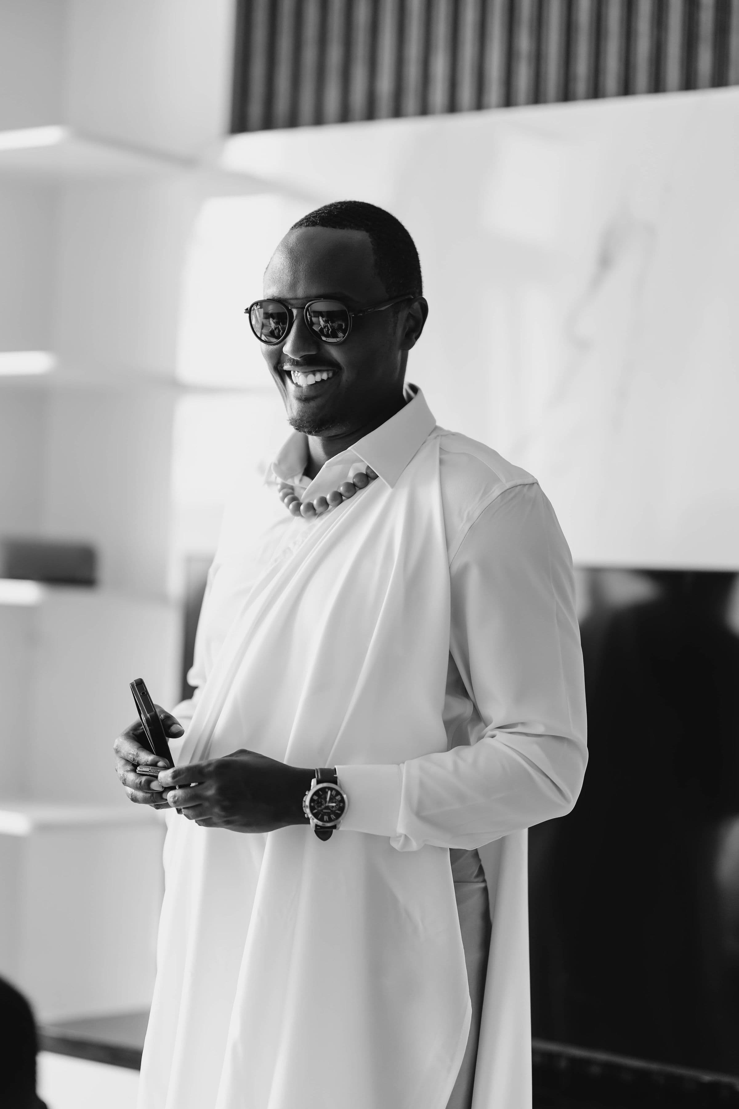 Mwanamume anayetabasamu akiwa amevaa miwani, vazi jeupe la kitamaduni, mkufu wa shanga na saa.