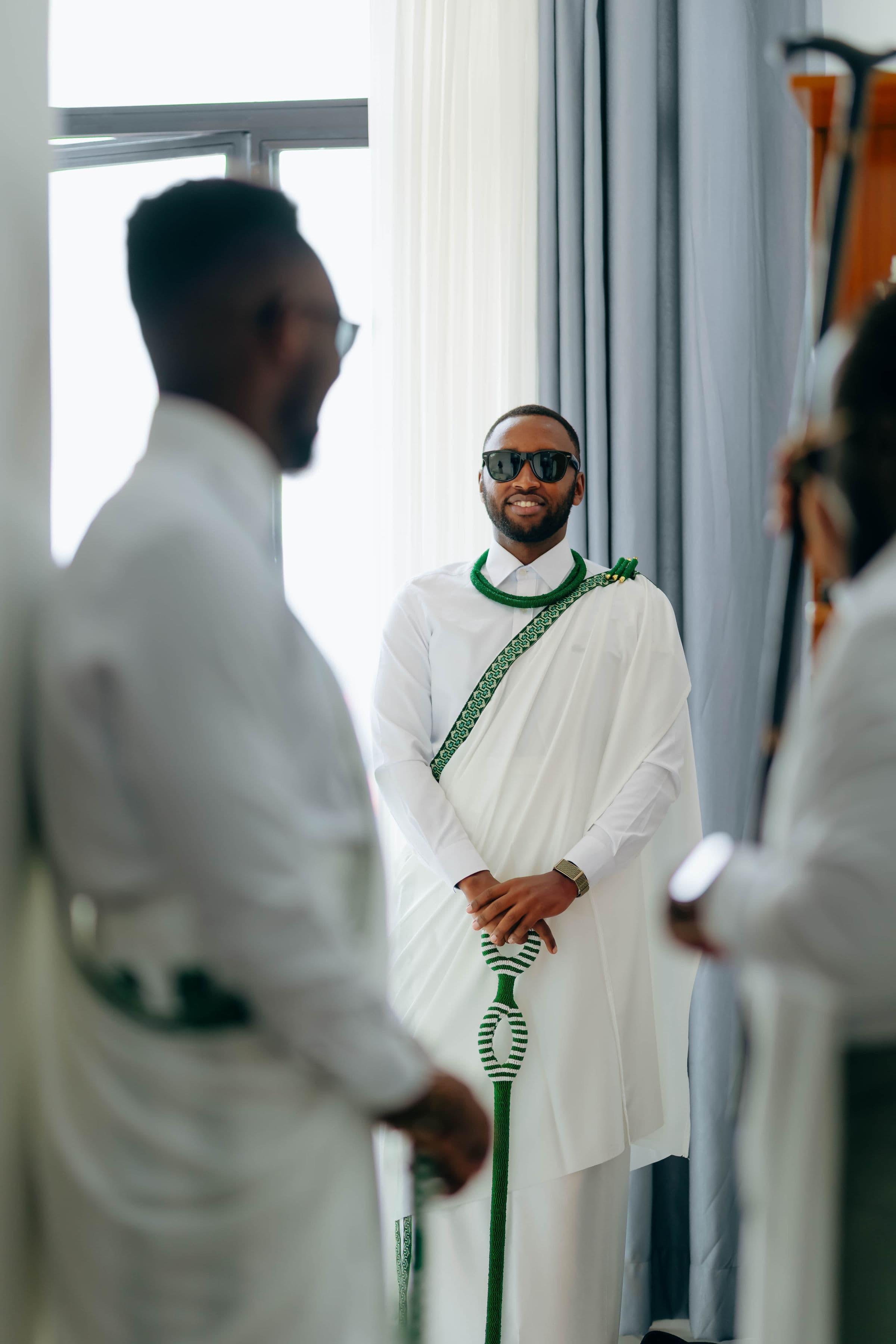 Mwanamume aliyevaa mavazi meupe ya kitamaduni ya Rwanda yenye shanga za kijani na miwani.