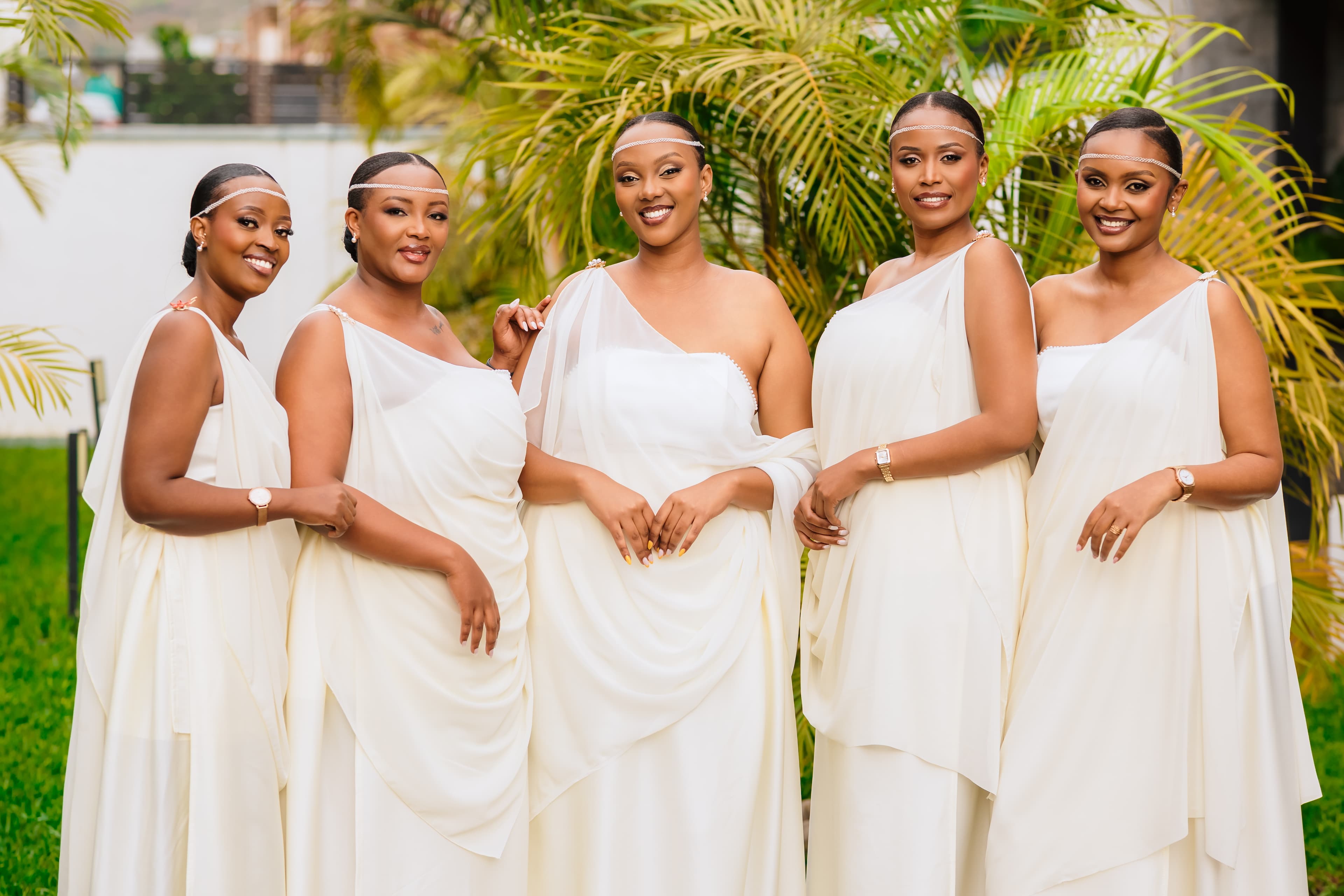 Wanawake watano wanaotabasamu wakiwa wamevalia mavazi meupe ya kitamaduni na vito vya kichwani.
