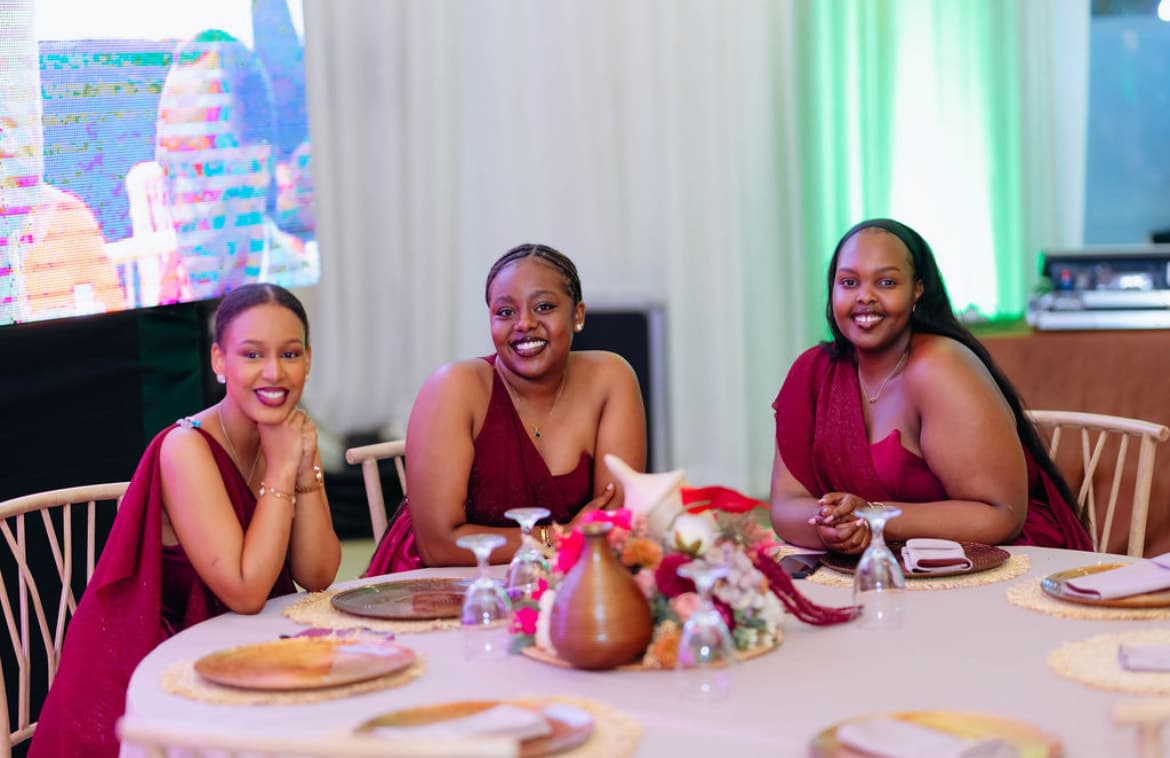 Wanawake watatu wanaotabasamu waliovalia mavazi ya maroon wameketi kwenye meza iliyopambwa wakati wa hafla.