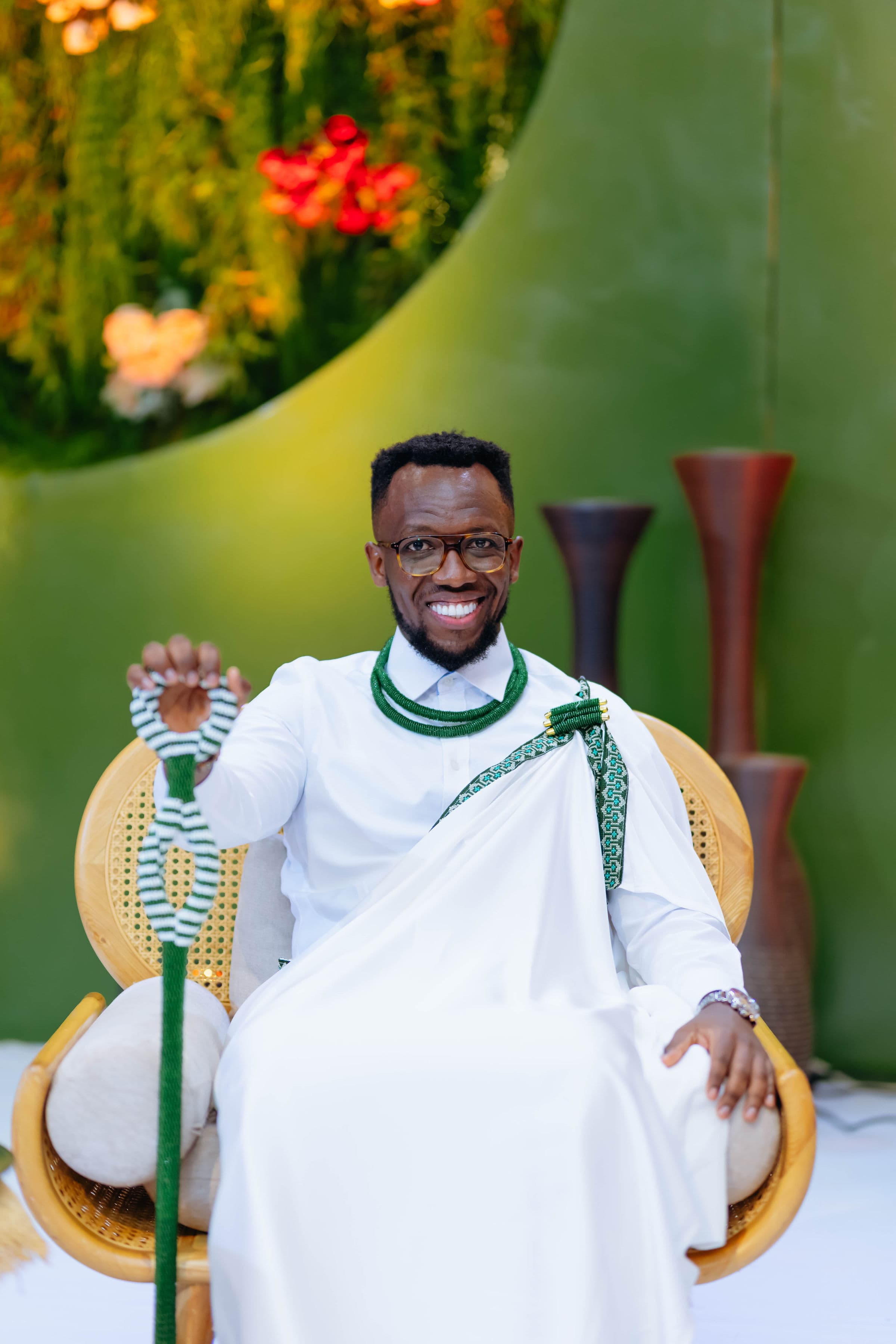 Mwanamume anayetabasamu akiwa amevaa mavazi meupe ya kitamaduni na shanga za kijani, ameketi akishika fimbo.